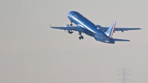 Ein blau lackiertes Verkehrsflugzeug hebt in den Himmel ab, während es leicht nach oben geneigt ist. Der Hintergrund ist neblig und zeigt leicht erkennbar eine Stromleitung in der Ferne.